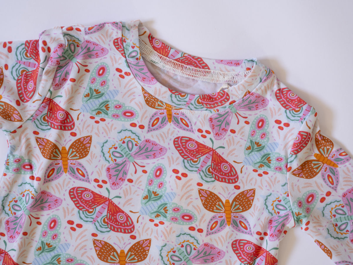 A long-sleeve baby top with a colorful butterfly and floral pattern on a white background, laid flat on a light surface.
