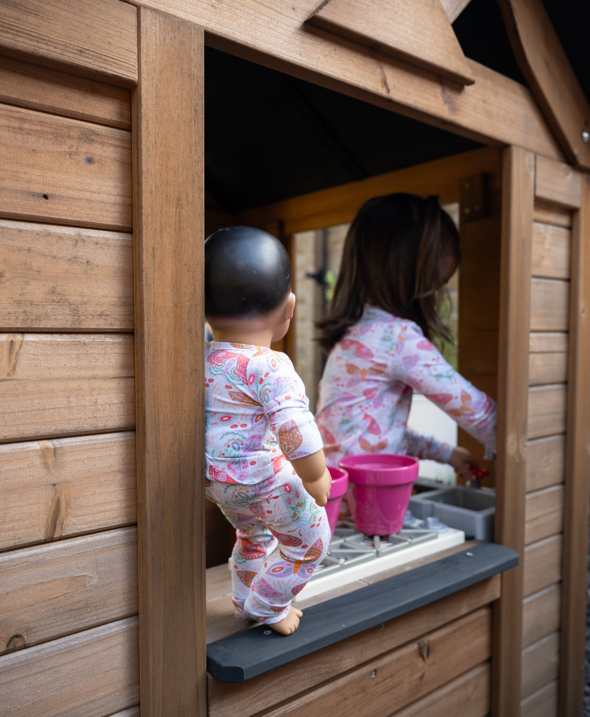 A baby in a pink floral onesie stands at the window of a wooden playhouse, looking into the play area where a child is playing with toy buckets and a small kitchen setup.