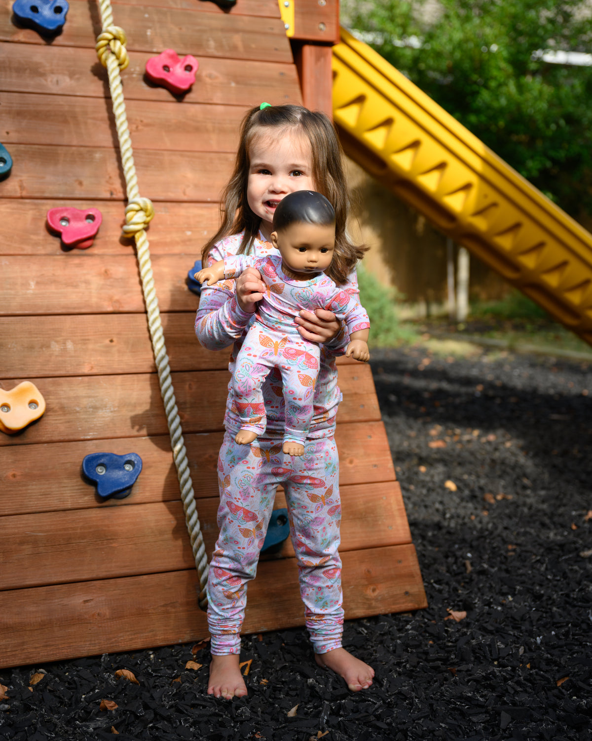 A young girl stands barefoot on a playground mulch surface in front of a wooden climbing wall, holding a baby doll. She is wearing a light pink and white patterned long-sleeve pajama top and matching pants.