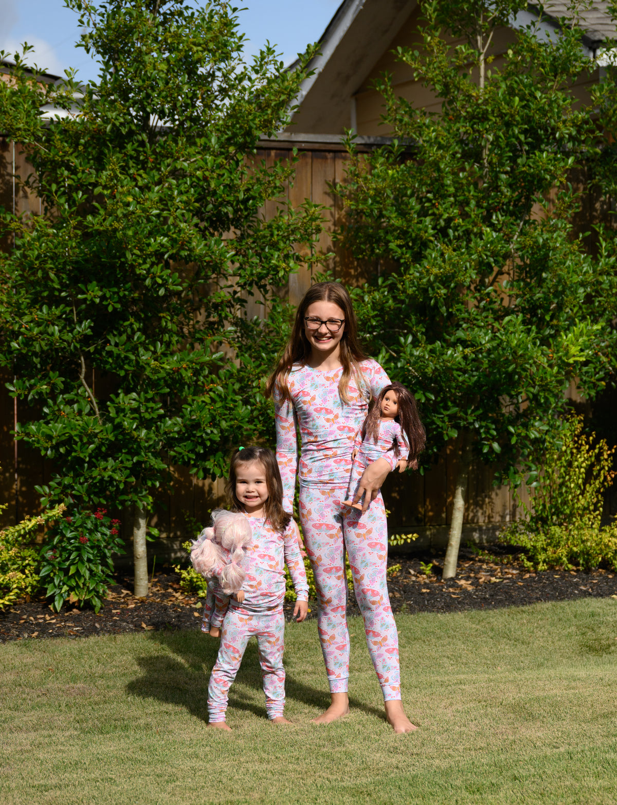 An older girl and a young girl wearing matching floral long-sleeve pajamas stand barefoot on a grassy lawn in front of a wooden fence and green trees. The both are holding dolls in matching pjs. animal.