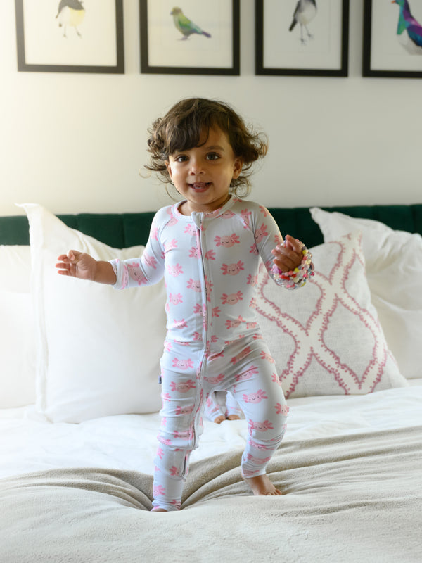 A toddler girl wearing a light pink and white footed pajama set with a pink axolotl pattern, standing on a white bed in a bedroom.